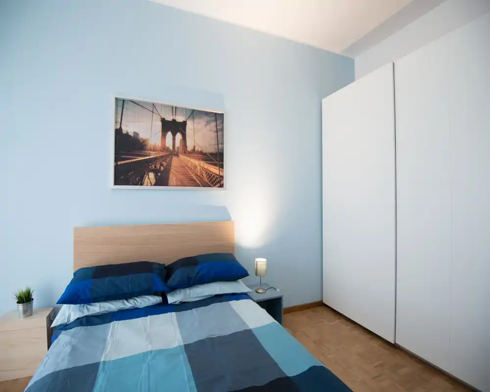 Dormitorio luminoso y ordenado con cama doble vestida con ropa azul de estilo geométrico, mesita con lámpara y gran armario. Las paredes azul claro y el cuadro crean un aire moderno y acogedor.