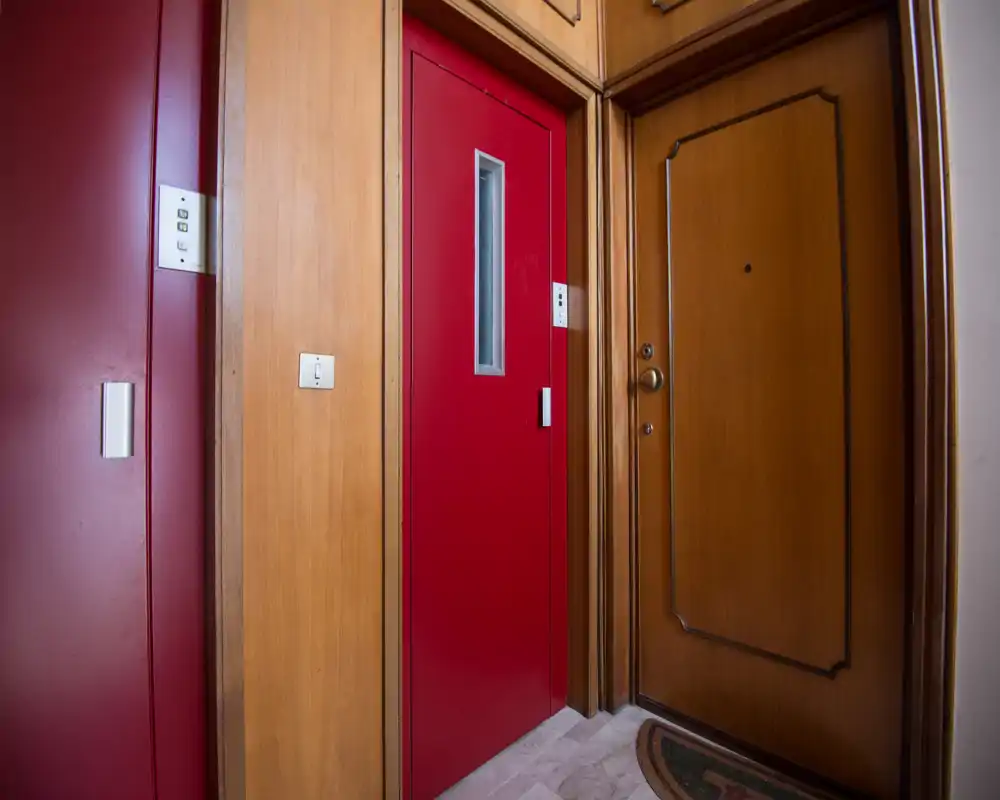 Entrada de edificio con puerta de madera del apartamento y puerta roja (ascensor/local); suelo de mármol y paneles de madera crean un recibidor ordenado y cuidado.