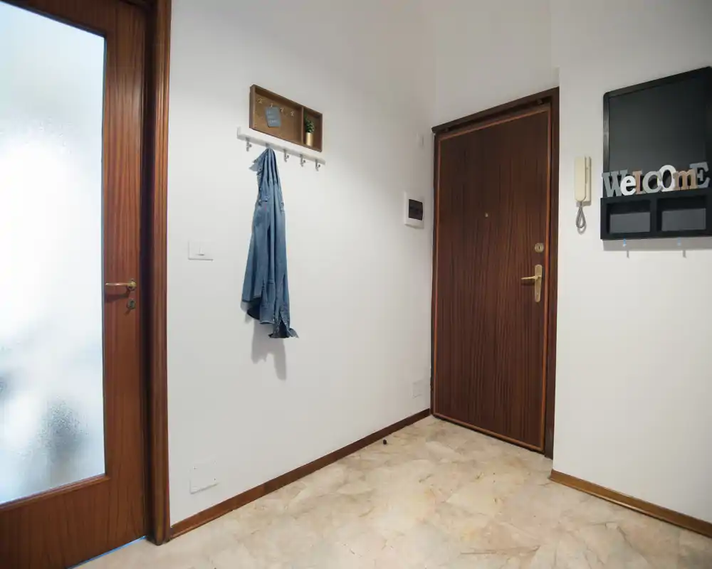 Entrada luminosa y ordenada con puerta principal de madera, perchas y puerta interior de vidrio esmerilado; suelo de mármol limpio y un organizador de pared sencillo.