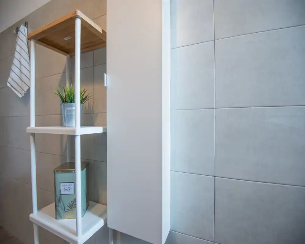 Rincón de baño minimalista con paredes alicatadas, un armario blanco estrecho y estanterías abiertas con pequeños objetos decorativos.