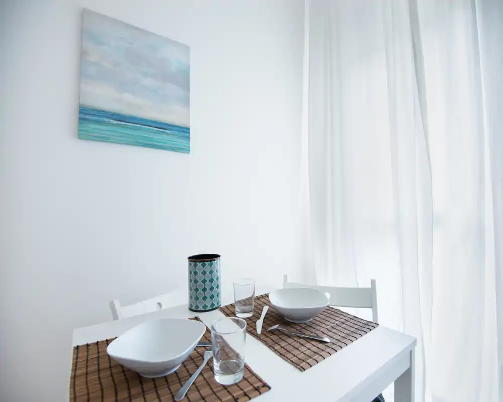 Pequeño rincón comedor luminoso con mesa blanca para dos, individuales texturizados, vajilla sencilla y luz natural suave a través de cortinas traslúcidas — un espacio minimalista y acogedor para comidas informales.