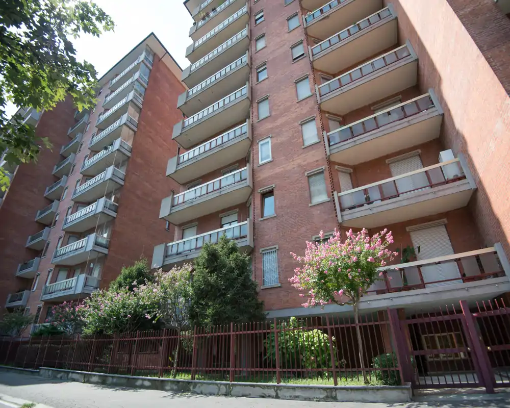 Vista exterior de un edificio residencial de ladrillo con balcones, reja frontal y arbustos en flor bajo luz diurna brillante.