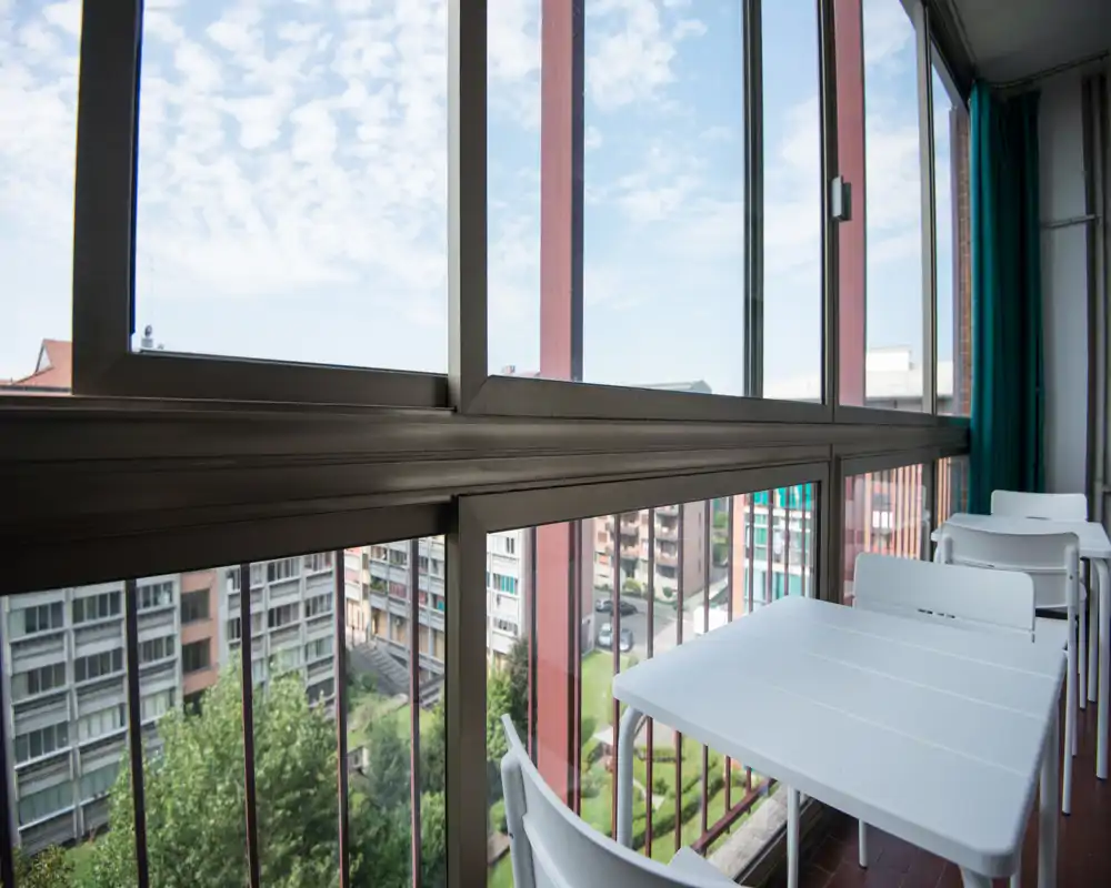 Balcón cerrado con grandes ventanas que dan a un patio residencial; mesa y sillas blancas sencillas crean un rincón acogedor para el café de la mañana con mucha luz natural.