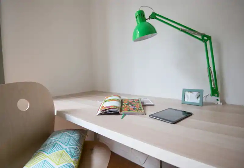 Bright minimal desk/working corner with a green adjustable lamp, a tablet and an open notebook on a light wood tabletop — a compact workspace ideal for studying or remote work.