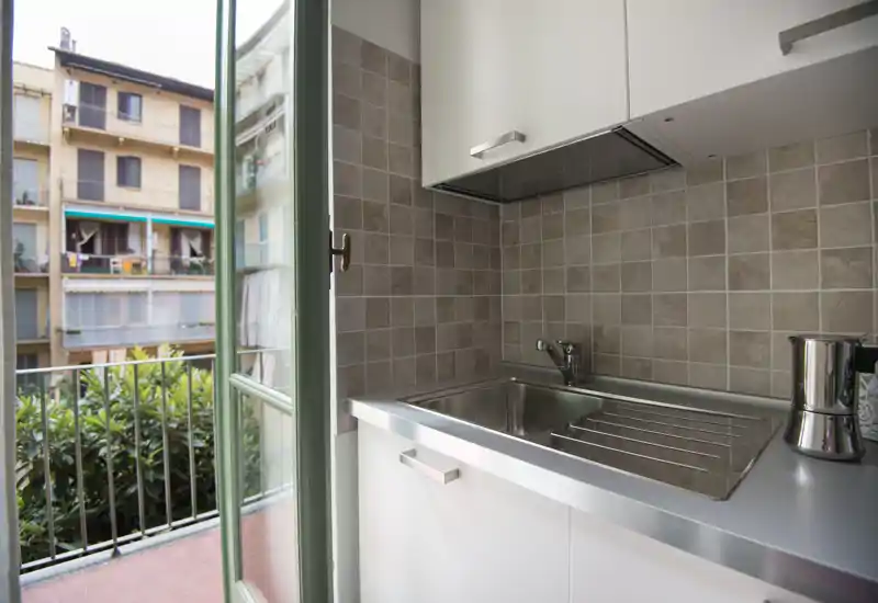 Bright compact kitchenette with stainless steel sink, tiled backsplash and an open balcony door providing natural light — ideal for a studio apartment.