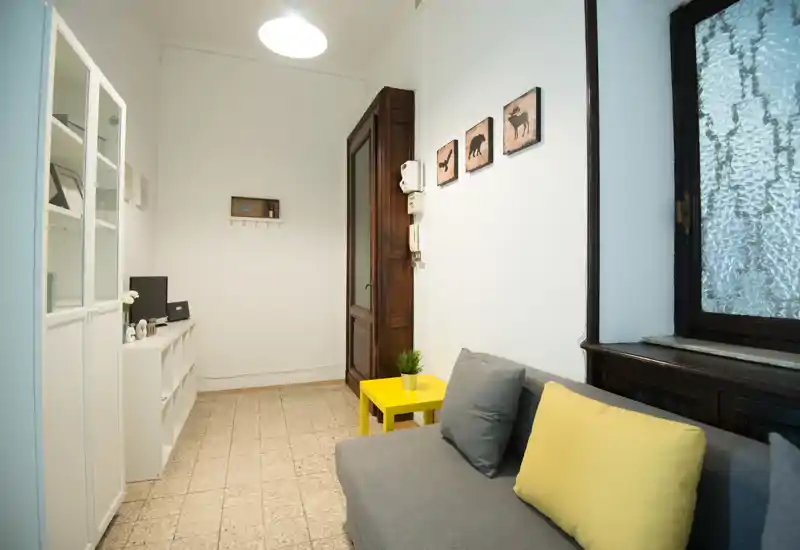 Compact, well-staged living area with a grey sofa, yellow accent pillows and a small yellow side table beside a tall wooden window frame. White storage cabinets and decorative wall art create a tidy, modern look.