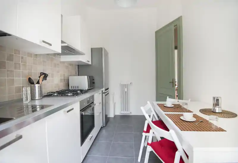 Bright, modern galley kitchen with stainless-steel appliances, tiled backsplash and a small dining table set for two with red seat cushions — clean and welcoming for short-term rentals.