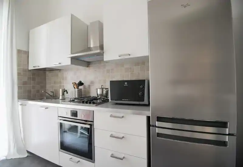 Modern compact kitchen with stainless-steel appliances, gas hob, built-in oven and tall refrigerator. Neutral tiled backsplash and white cabinetry create a bright, clean cooking area.