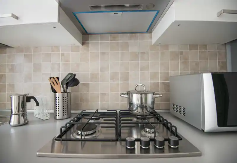 Modern compact kitchen with a stainless steel gas cooktop, utensils holder, moka pot and a microwave on the countertop; neutral tiled backsplash and clean, minimalist surfaces.
