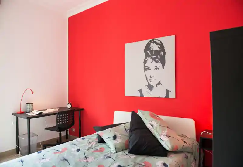 Dormitorio luminoso y moderno con edredón estampado, cojines decorativos y una pared roja llamativa; cuenta con escritorio y mesita de noche.
