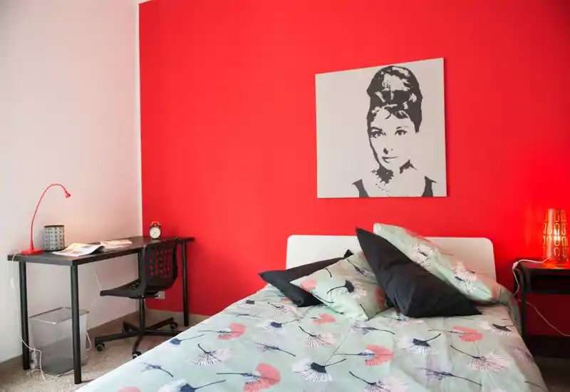 Dormitorio luminoso y con estilo con colcha estampada, cojines decorativos y pared roja de acento; incluye un pequeño escritorio y lámpara de mesilla, ideal para un anuncio acogedor.