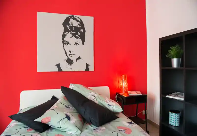 Dormitorio luminoso y con estilo, con ropa de cama estampada, cojines decorativos y una pared roja llamativa con gran cuadro; mesita con lámpara y estantería que completan el ambiente acogedor.