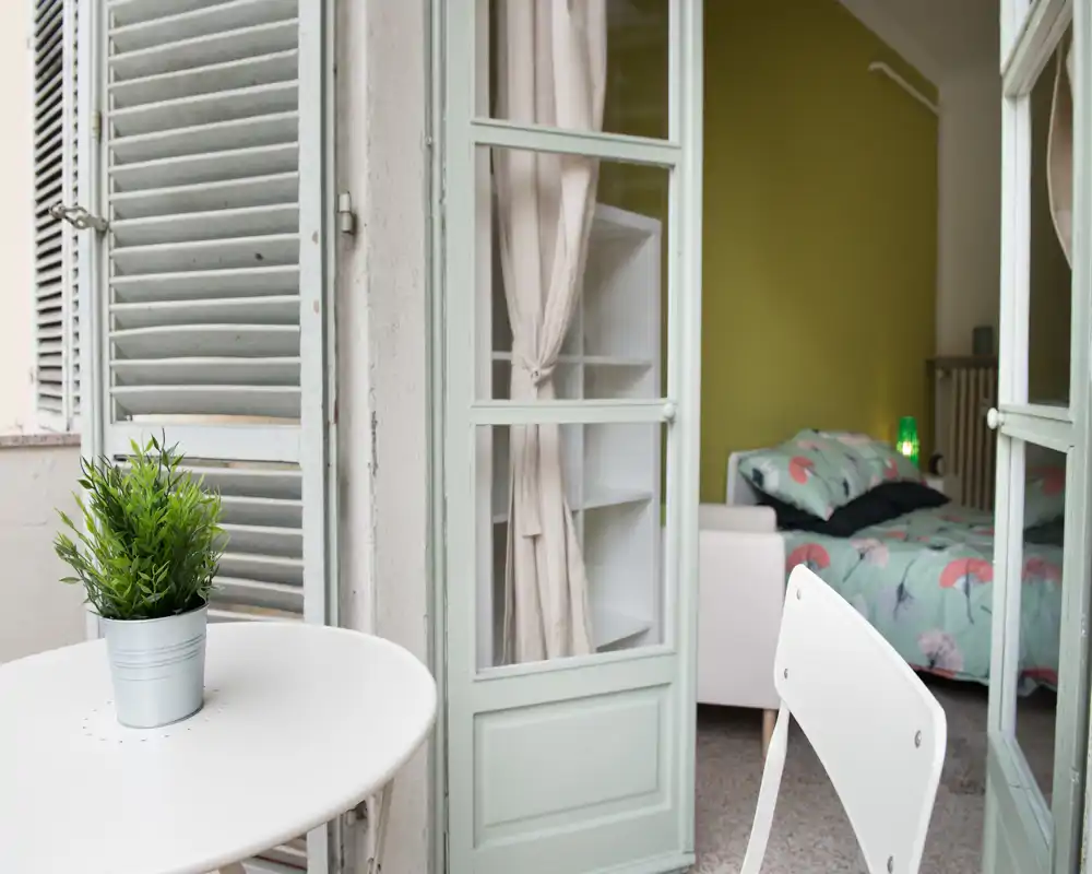 Sunny bedroom seen through French doors opening to a small balcony; patterned bedding, bedside lamp and soft natural light create a cozy, well-staged sleeping area.