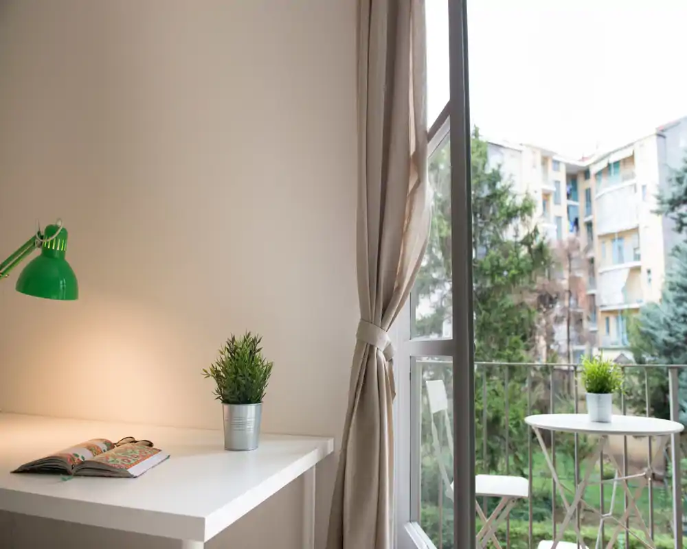 A small balcony with a bistro table and chairs seen through a glass door; a potted plant on the table and green views outside create a pleasant outdoor spot.