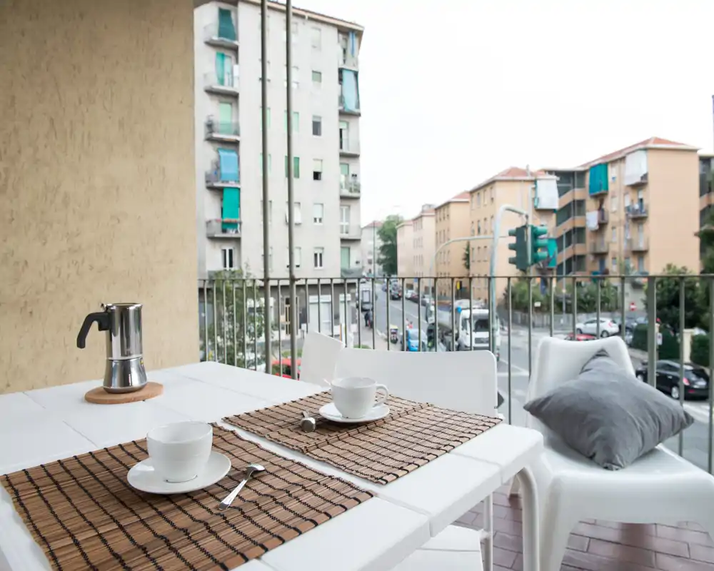 Balcón soleado con pequeño comedor: mesa y sillas blancas con manteles, tazas y cafetera moka, con vistas a una calle urbana tranquila.