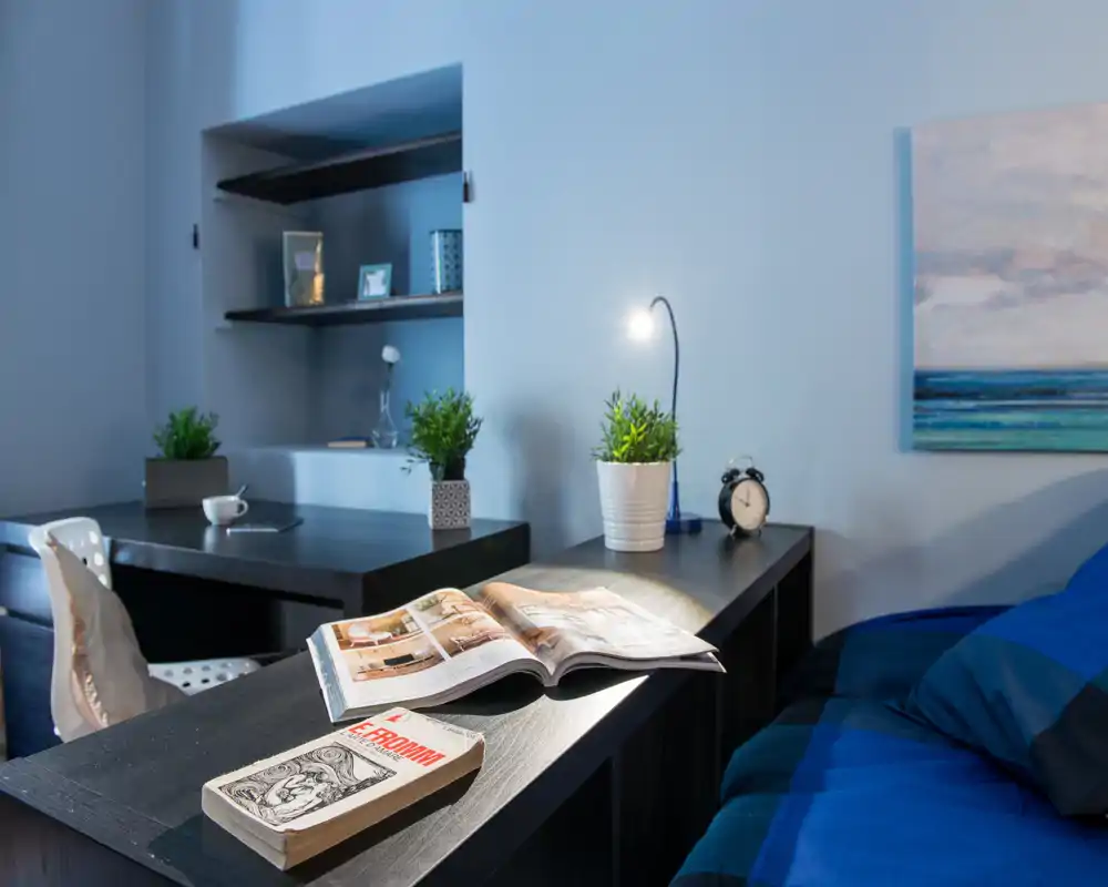 Rincón de dormitorio ambientado con cama visible con ropa de cama azul, mesita con lámpara, plantas, libros y zona de trabajo—luminoso, ordenado y bien compuesto para un anuncio.