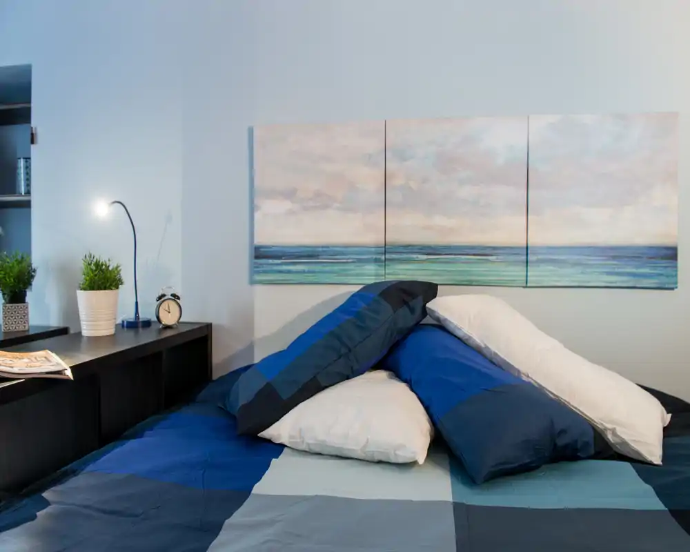 Dormitorio luminoso y bien decorado con colcha azul, varias almohadas y un cuadro de mar sobre la cama. La estantería de la mesilla con lámpara, reloj y plantas aporta un ambiente acogedor y moderno.