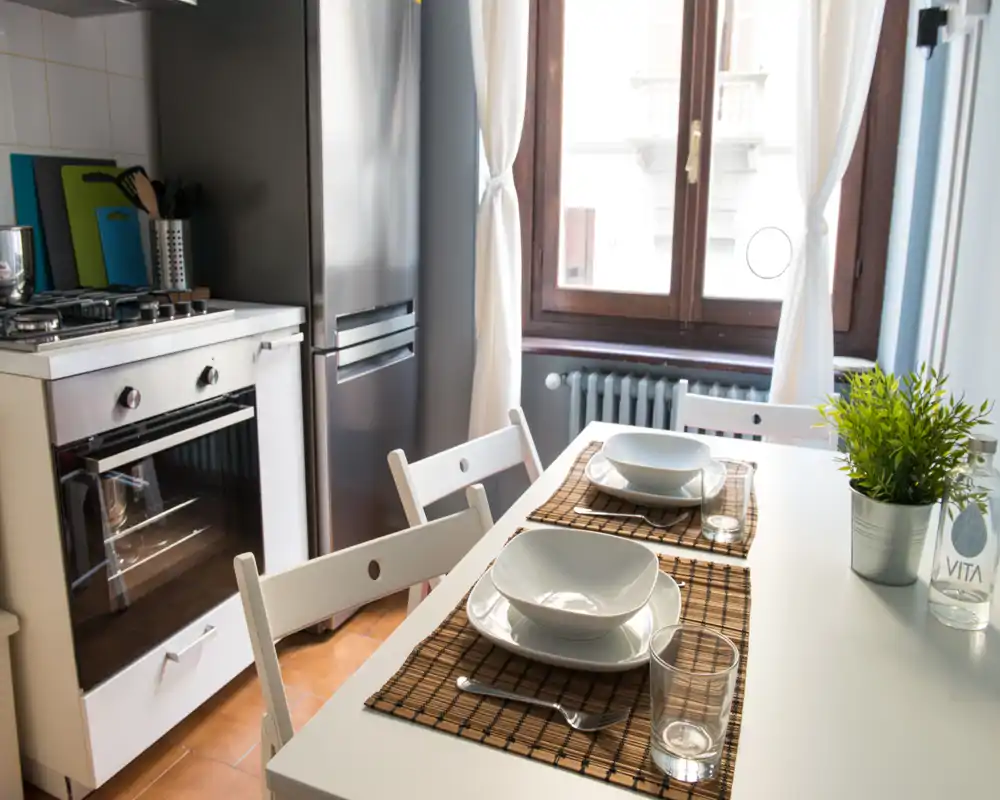 Cocina moderna y luminosa con mesa puesta para dos, electrodomésticos de acero inoxidable y una gran ventana que aporta luz natural.