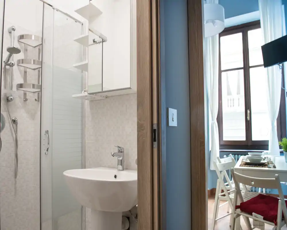 Baño compacto con lavabo de pie y ducha con mampara de cristal, fotografiado junto a una puerta que da a un luminoso comedor.