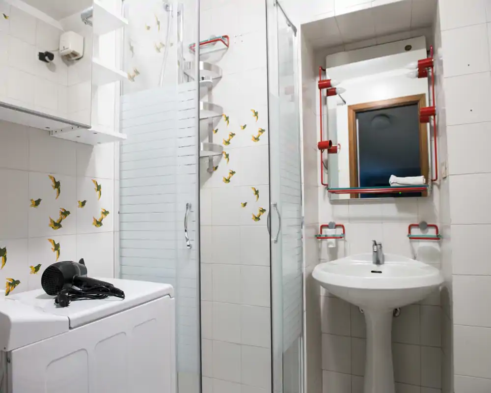 Baño compacto y bien iluminado con lavabo de pie, espejo, mampara de ducha de vidrio y azulejos con motivos decorativos.