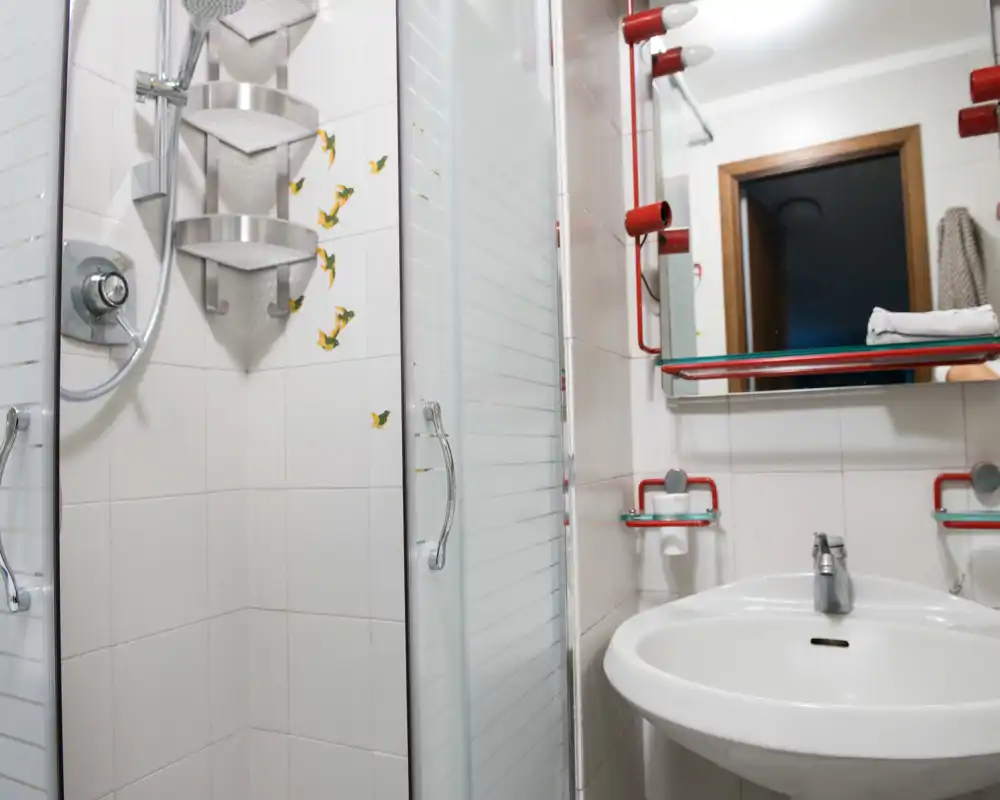 Baño pequeño y luminoso con ducha de esquina de cristal, estantería de pared, lavabo con espejo y grifería moderna.