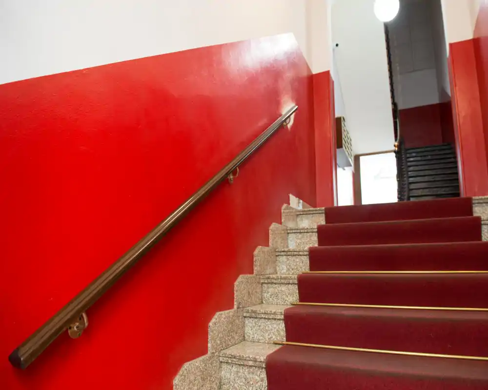 Escalera luminosa y bien cuidada con paredes rojas, pasamanos de madera y escalones con alfombra roja — foto ideal para mostrar la entrada y las zonas comunes.