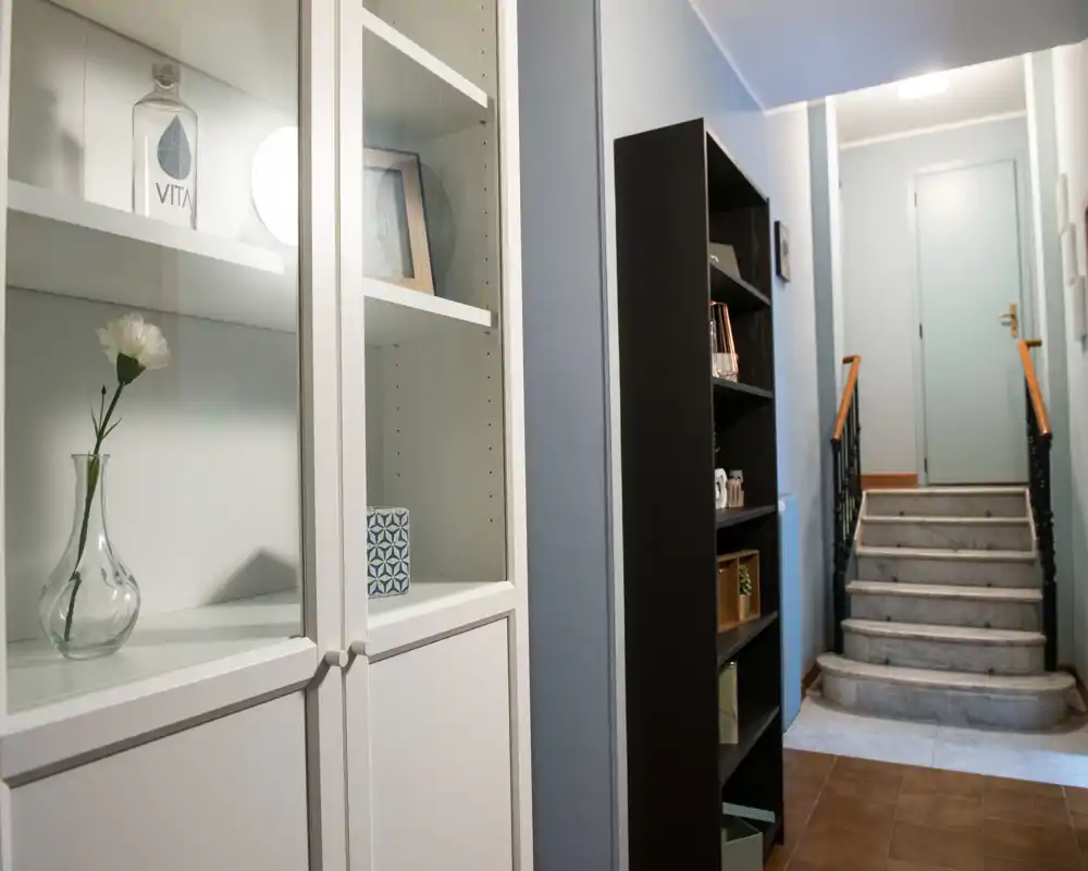 Recibidor bien iluminado con estanterías decorativas y una pequeña escalera que conduce a una puerta interior — ordenado, moderno y acogedor.