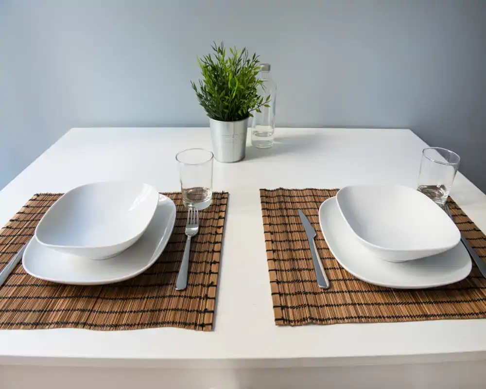 Mesa minimalista para dos con platos blancos cuadrados, cristalería y una pequeña planta en maceta sobre una mesa blanca y luminosa — moderna, ordenada y lista para una comida informal.
