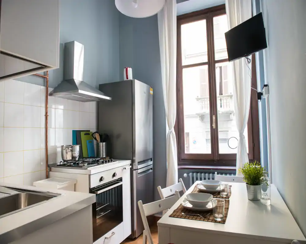 Cocina compacta y luminosa con vitrocerámica a gas, campana y frigorífico junto a un rincón comedor junto a la ventana, práctica y acogedora para estancias cortas.