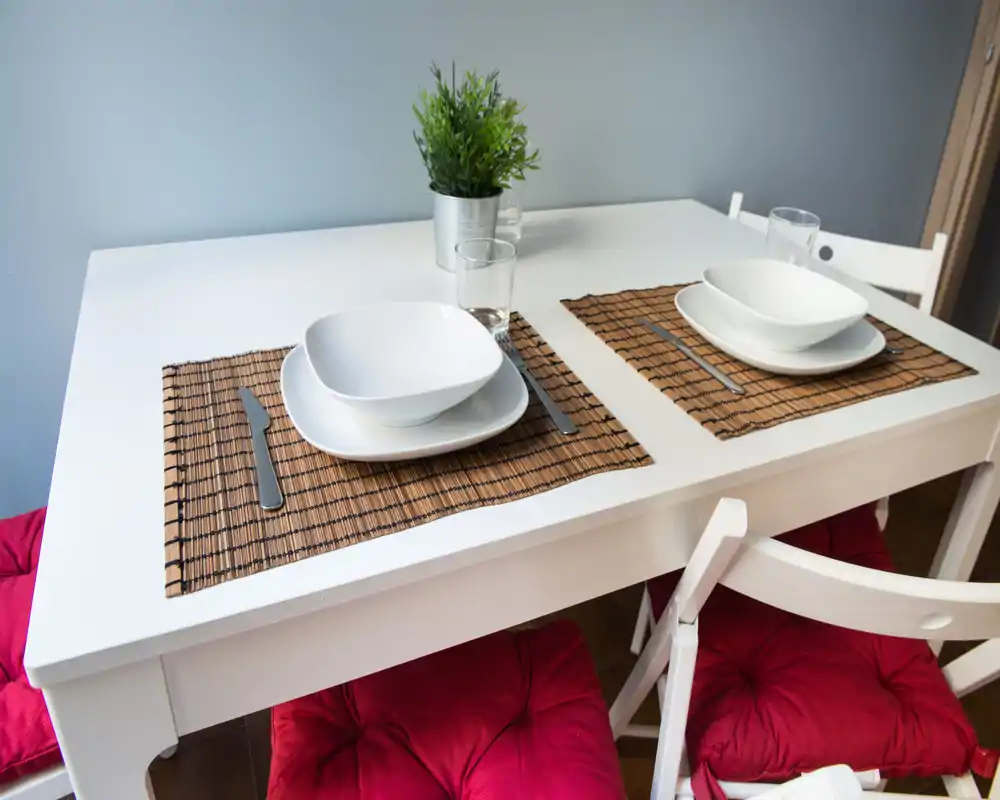 Mesa de comedor para dos ordenada, con individuales de mimbre, cuencos blancos modernos y sillas con cojines rojos; brillante, minimalista y bien ambientada para un área de comedor compacta.