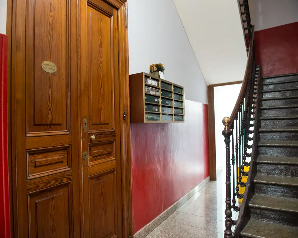 Entrada de edificio bien iluminada con puerta de madera, buzones en la pared y una barandilla decorativa en la escalera; suelo de piedra pulida y pasillo luminoso.