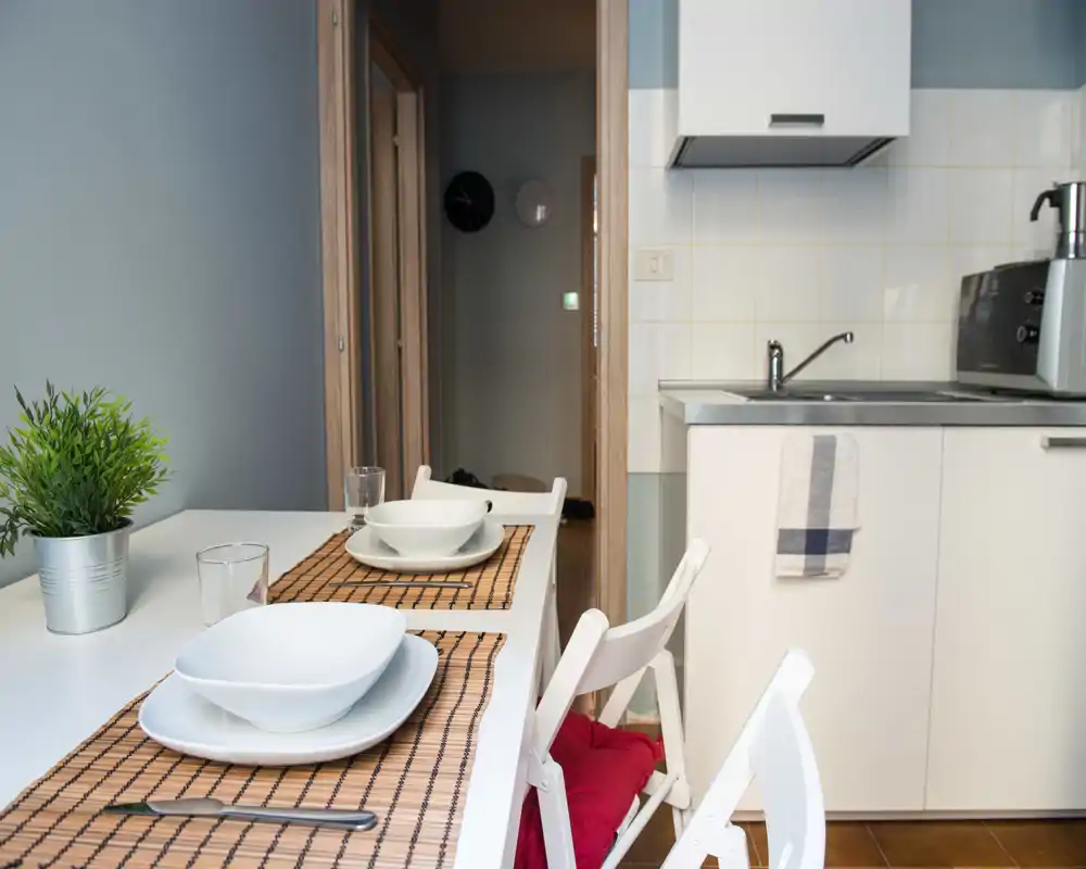 Cocina compacta con zona de comedor: muebles blancos, fregadero de acero, mesa puesta para dos con individuales y cuencos, y una planta en maceta que aporta verdor.