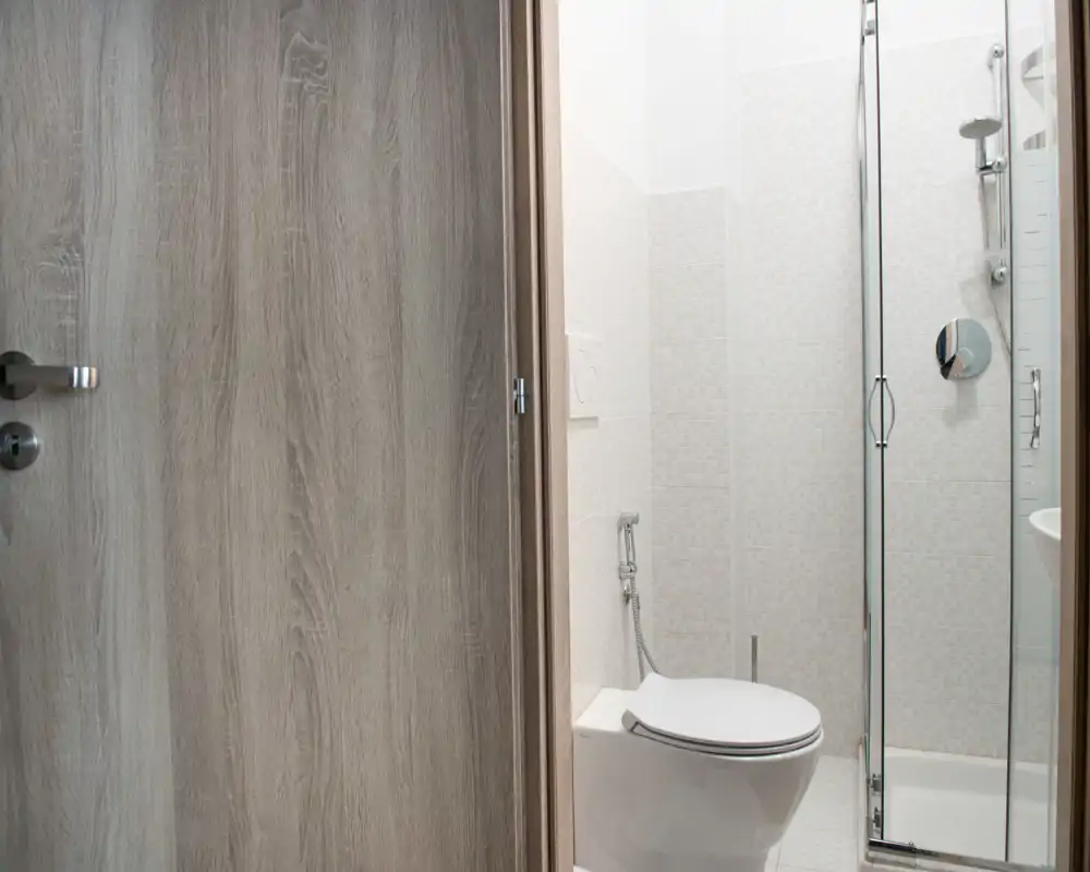 Baño moderno y compacto con inodoro suspendido y ducha con mampara de cristal, azulejos claros y grifería contemporánea.
