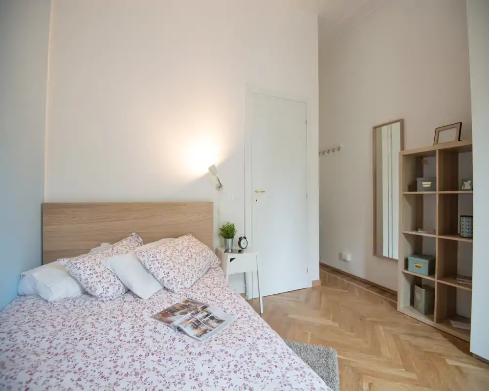 Camera da letto luminosa e ordinata con copripiumino a fantasia, testiera in legno e comodino con lampada e pianta; pavimento in parquet e mensole minimali per un ambiente rilassante.