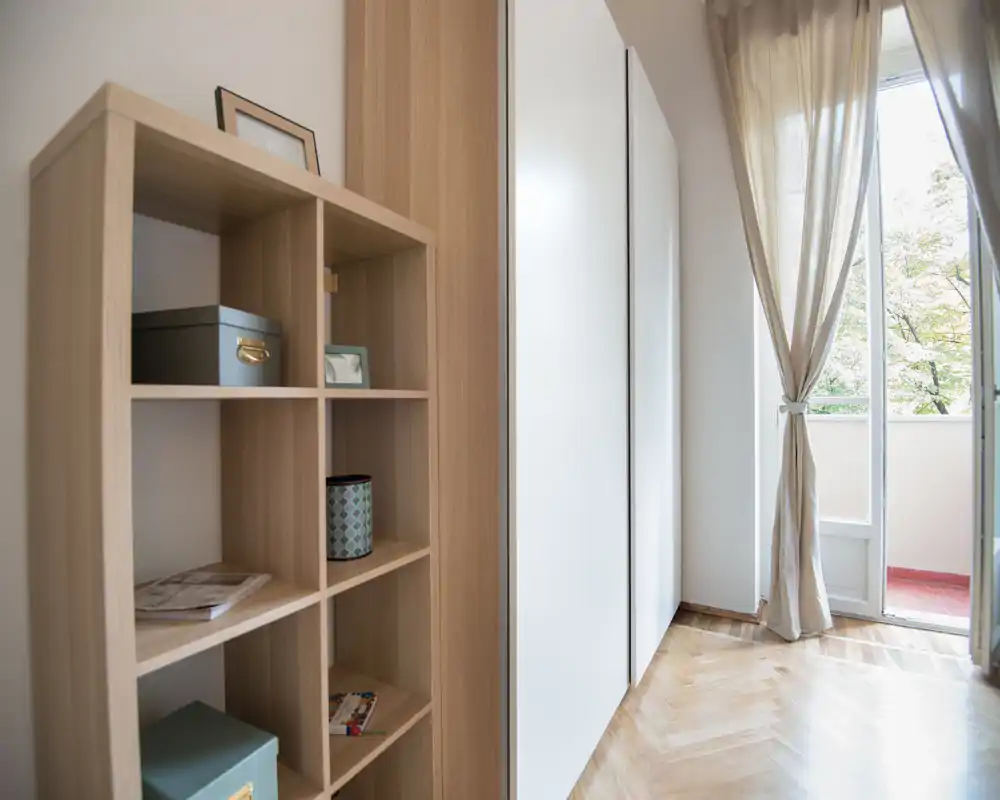 Angolo luminoso e minimale del soggiorno con scaffalatura in legno, armadi a muro e una porta con tenda leggera che si apre su un piccolo balcone — pulito e arioso.