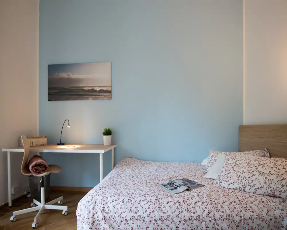Camera da letto luminosa e accogliente con biancheria a fantasia, testiera in legno e una piccola scrivania con lampada — perfetta per riposo o smart working.