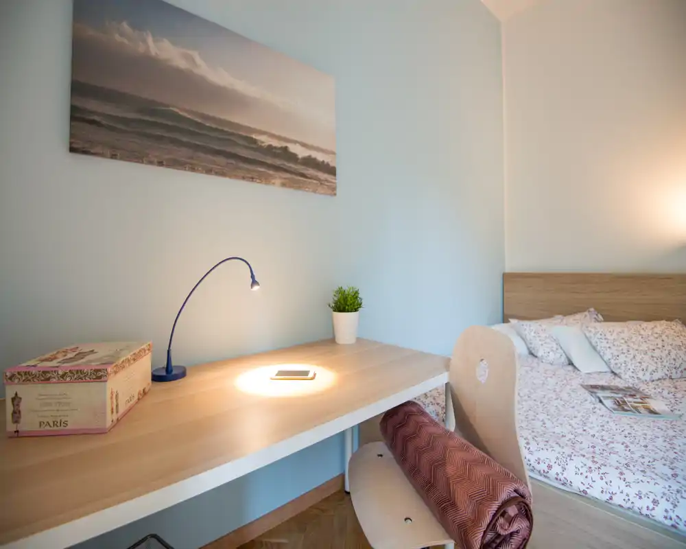 Angolo camera luminoso e curato con scrivania in legno e letto ben rifatto con lenzuola floreali; lampada da tavolo calda e una piccola pianta rendono l'ambiente accogliente.