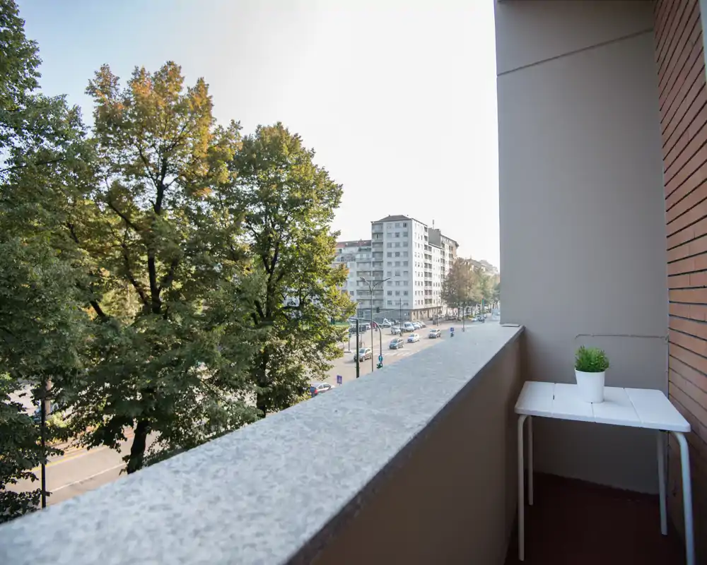 Balcone soleggiato con vista su una strada alberata, piccolo tavolo e vaso di pianta — perfetto per il caffè mattutino e la vista sulla città.