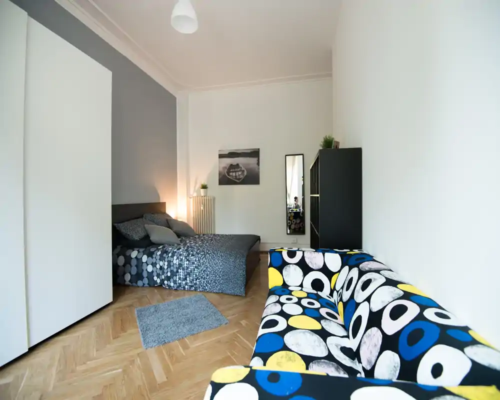 Dormitorio luminoso y con estilo que incluye una cama doble con ropa de cama estampada, un sofá colorido en primer plano, armario y cuadros en la pared. Suelo de madera y luz ambiental suave crean un ambiente acogedor.