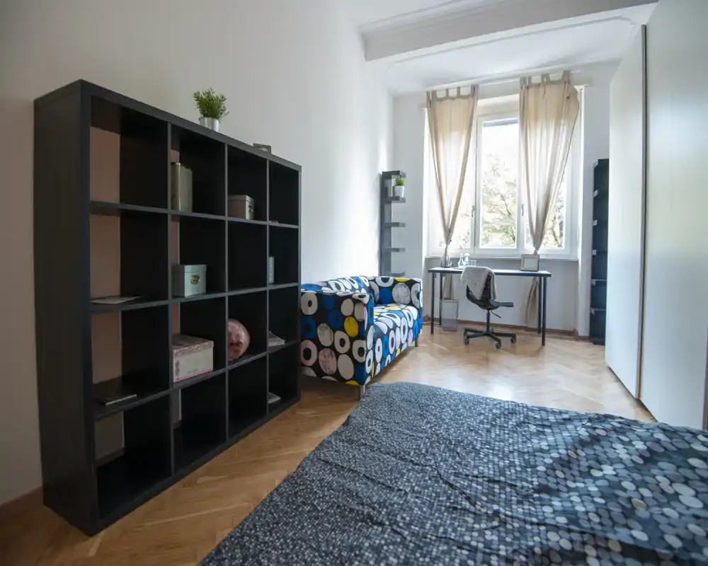 Dormitorio luminoso y moderno con un sofá estampado, escritorio junto a la ventana y la cama visible en primer plano. Suelo de madera y paredes neutras crean un ambiente limpio y luminoso, ideal para anuncios.