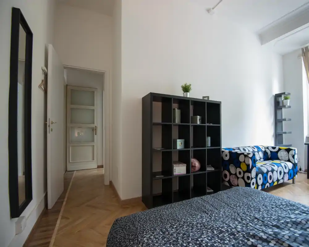 Rincón de dormitorio moderno y luminoso con un sofá estampado, estanterías abiertas y cama parcialmente visible en primer plano. La luz natural y la decoración minimalista crean un ambiente acogedor, perfecto para alquileres a corto plazo.