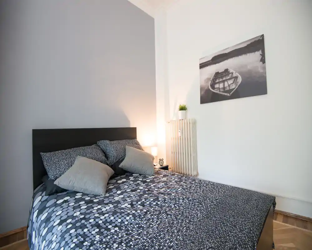 Dormitorio moderno y luminoso con cama doble, ropa de cama estampada, cojines decorativos, lámpara de mesita y cuadro en la pared — ideal para una estancia confortable.