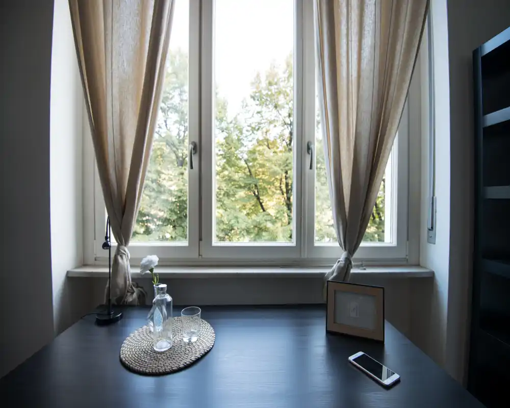 Rincón junto a la ventana luminoso con mesa oscura, decoración sencilla y foto enmarcada: un tranquilo espacio de estar/trabajo con luz natural.