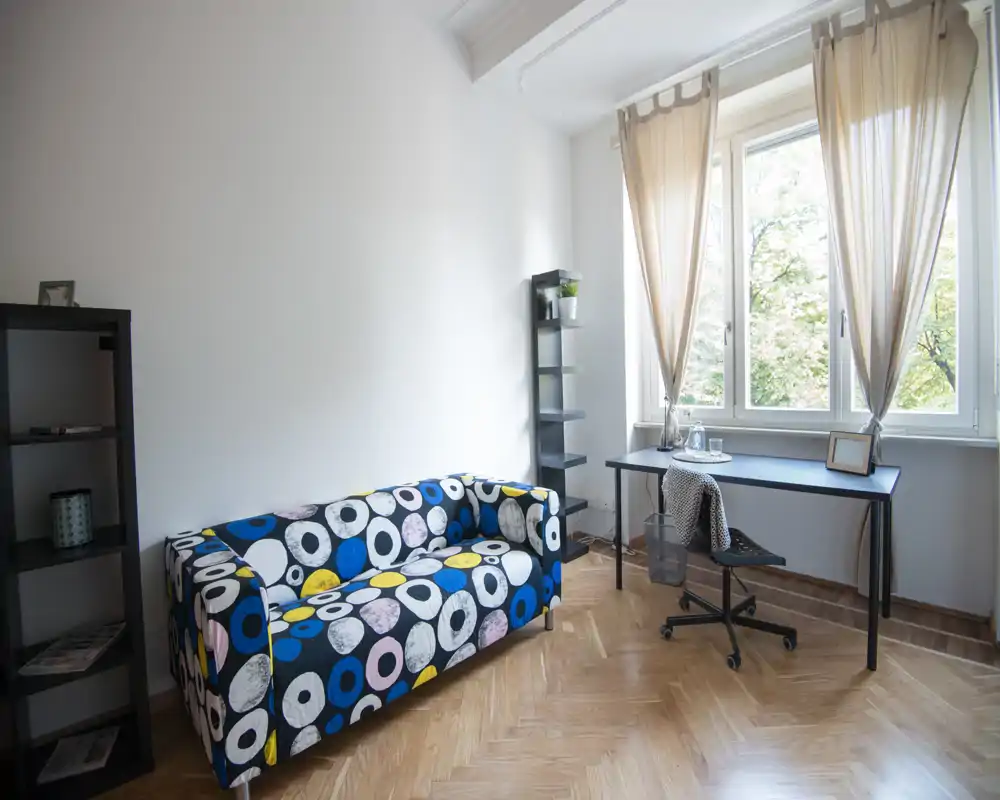 Zona de estar luminosa y moderna con un sofá colorido estampado, un escritorio junto a la ventana y estanterías abiertas — ideal para un estudio o un rincón de teletrabajo.