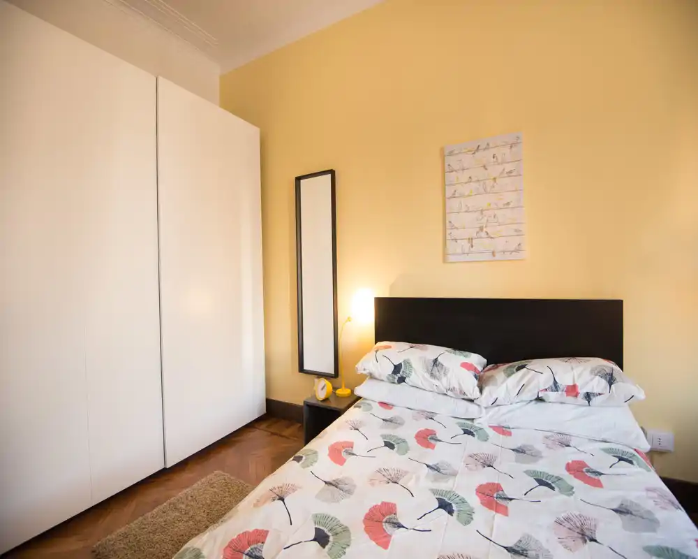 Cozy, well-styled bedroom with a double bed, floral-patterned bedding, bedside lamp and a large wardrobe — warm yellow walls create a welcoming atmosphere.