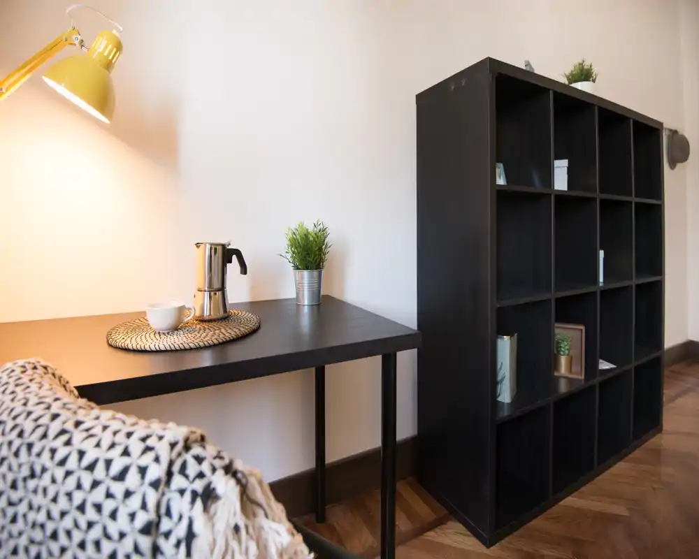 Bright, stylish living corner with a black shelving unit, minimalist desk with lamp and decorative plants — cozy and well-styled for a short-term rental.