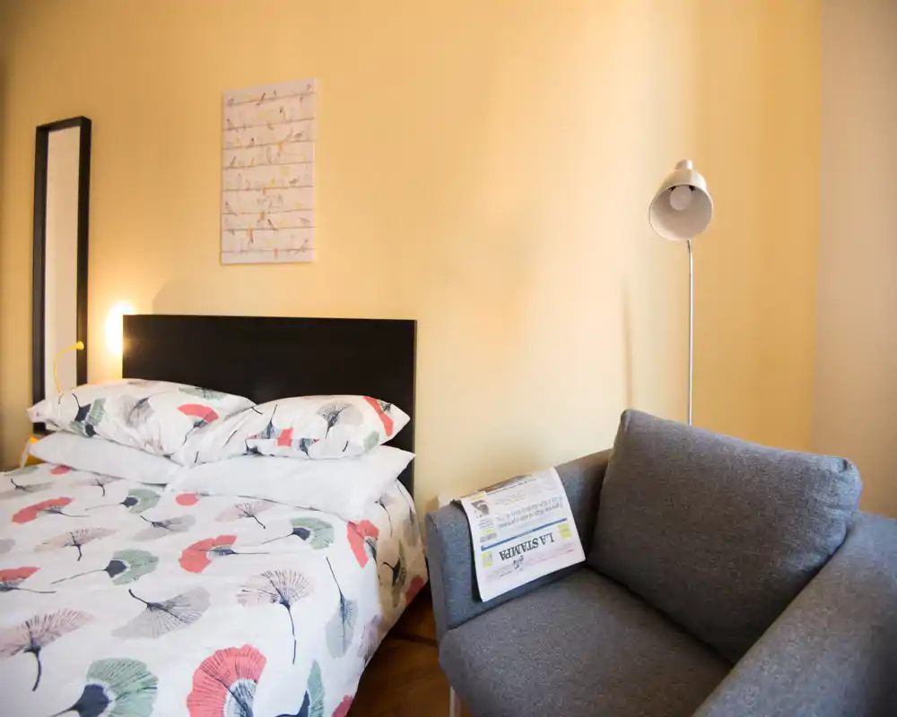 Bright, cozy bedroom with a patterned double bed, dark headboard and a small gray sofa beside it — warm yellow walls and a standing lamp create a welcoming atmosphere.