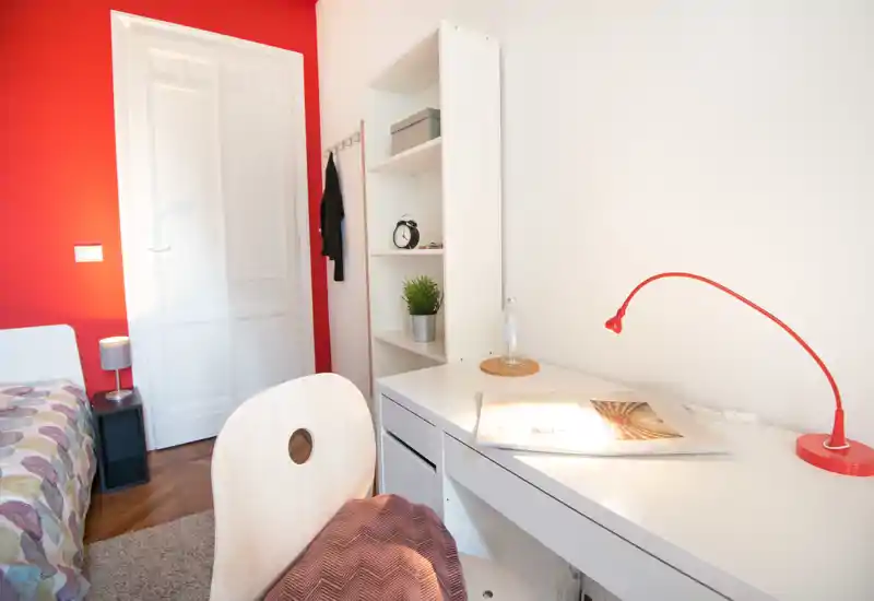 Coin chambre lumineux et moderne avec bureau blanc, lampe rouge et partie d'un lit à motif. Une étagère blanche et un mur rouge apportent du caractère.