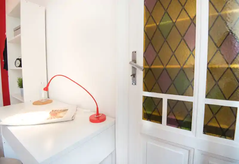Bright, minimal interior corner with a white desk, red reading lamp and decorative shelf beside a door with stained-glass panes — a tidy living/corner workspace with natural light.