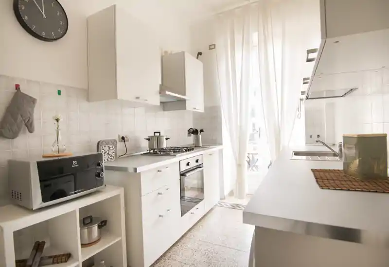 Bright modern kitchen with white cabinetry, gas stove and oven, microwave and sink illuminated by natural light from a tall window/door; clean, minimal styling ideal for rental listings.